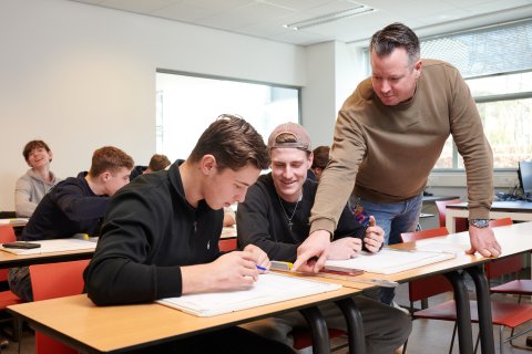 Docent staat naast de tafel van twee studenten en wijst iets aan op de bouwtekening die de student heeft gemaakt