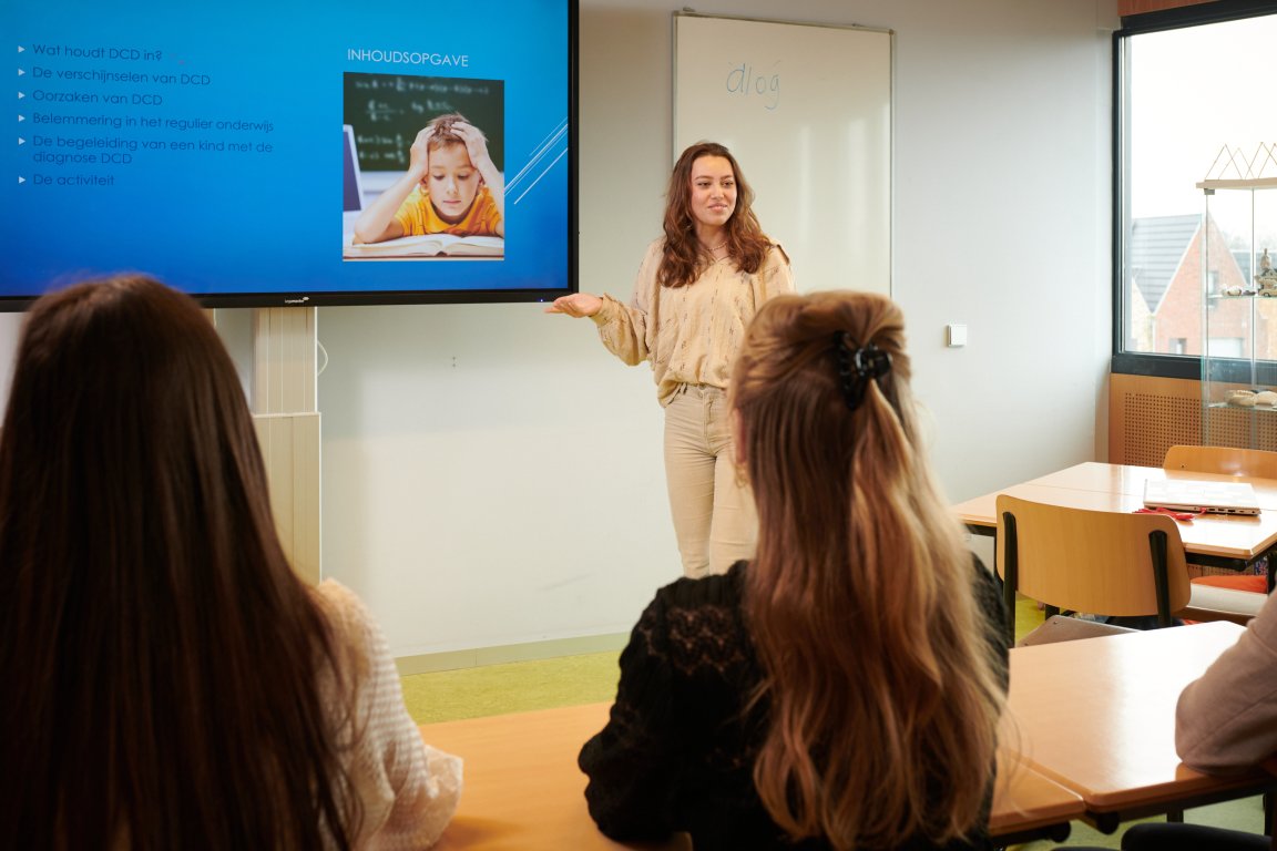 Docent staat voor de klas naast een groot scherm waarop een presentatie afspeelt over onderwijs aan kinderen