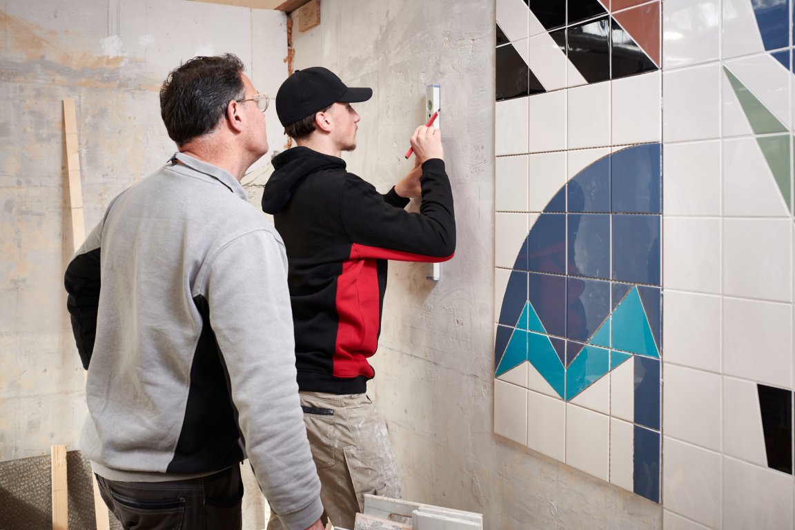 Docent kijkt mee met student die met een waterpas en potlood op de muur een streep zet naast een stuk muur dat betegeld is