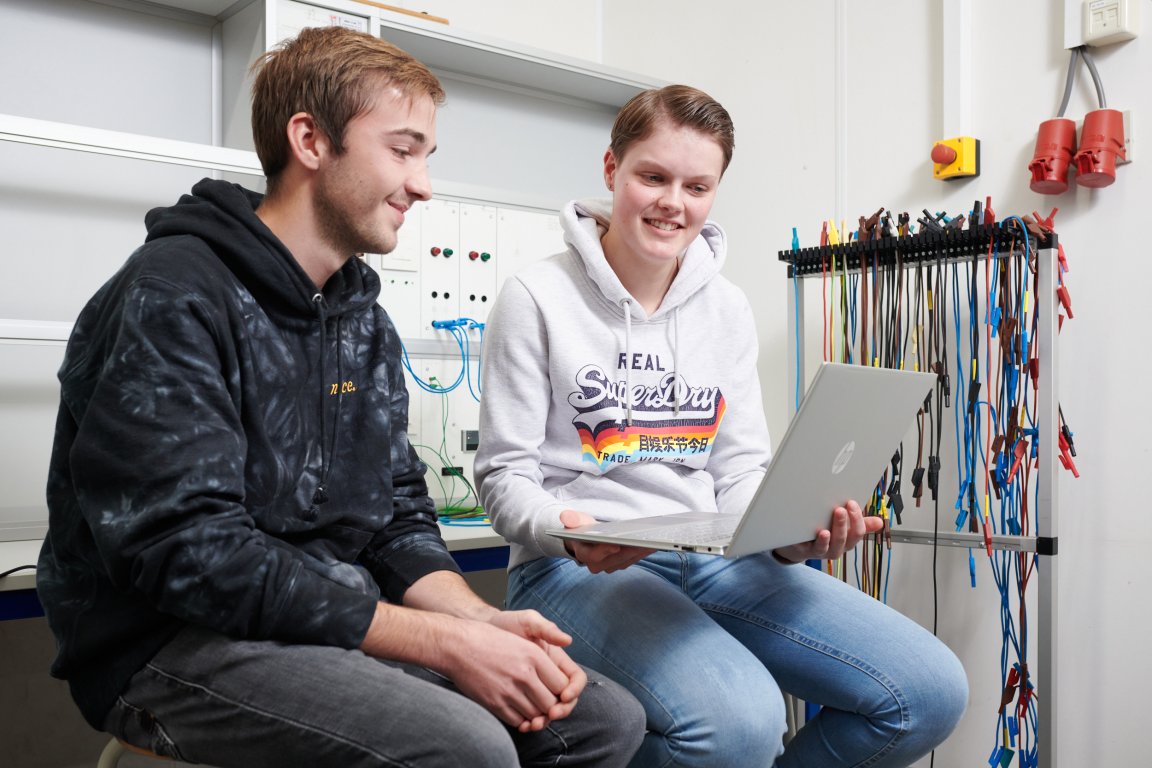 Twee studenten zittend op tafel waarvan één van hen een laptop vasthoud. Achter hen is een hoop bedrading te zien.