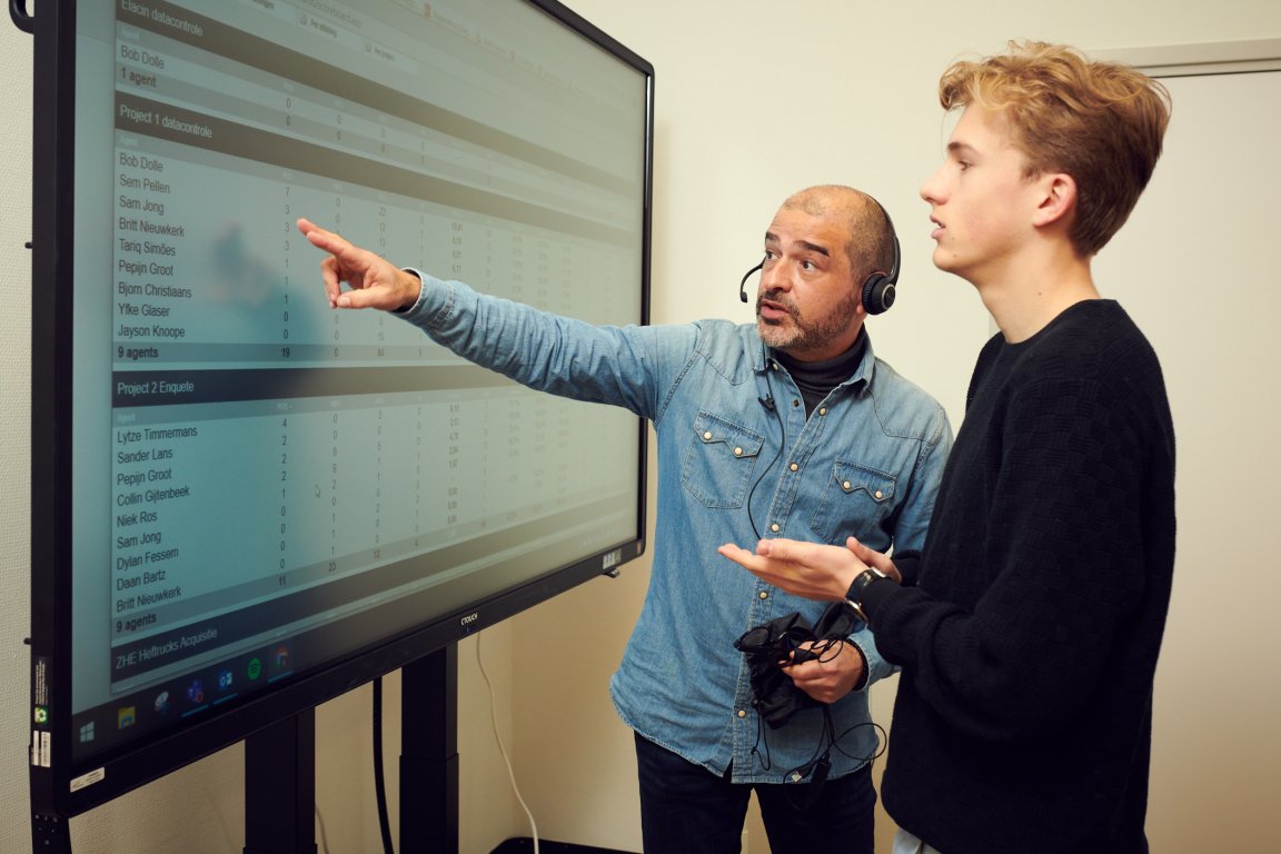 Docent met headset op wijst zijn student iets aan op het scherm