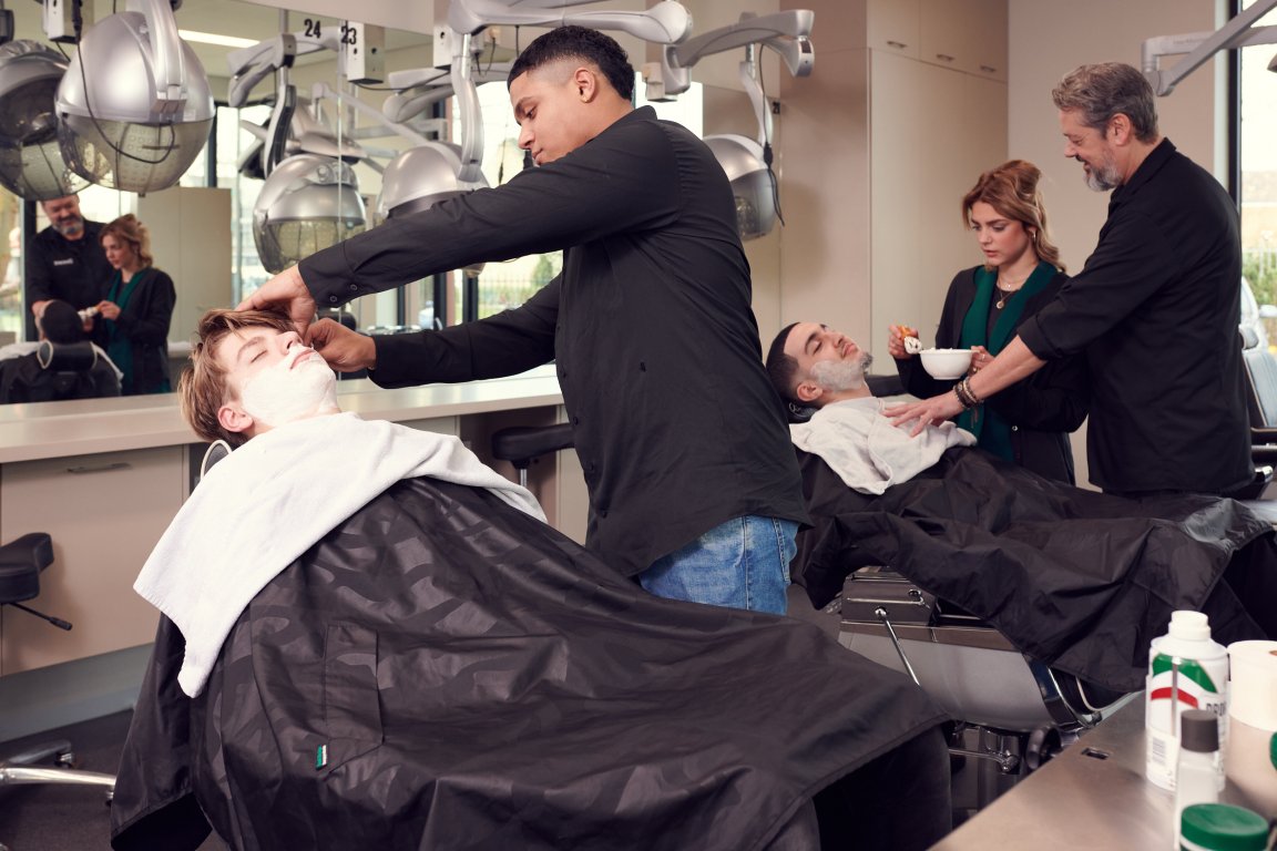 Twee studenten oefenen op medestudent het trimmen van de baard van een heer. Een docent staat erbij en geeft aanwijzingen.