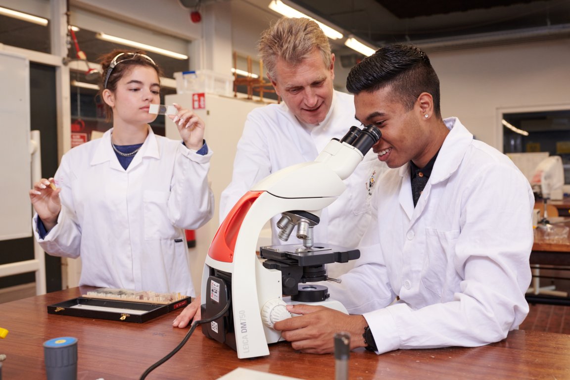 Student kijkt door een microscoop terwijl een docent ernaast aanwijzingen geeft en een andere student bekijkt het glaasje met het monster dat ze gaan onderzoeken