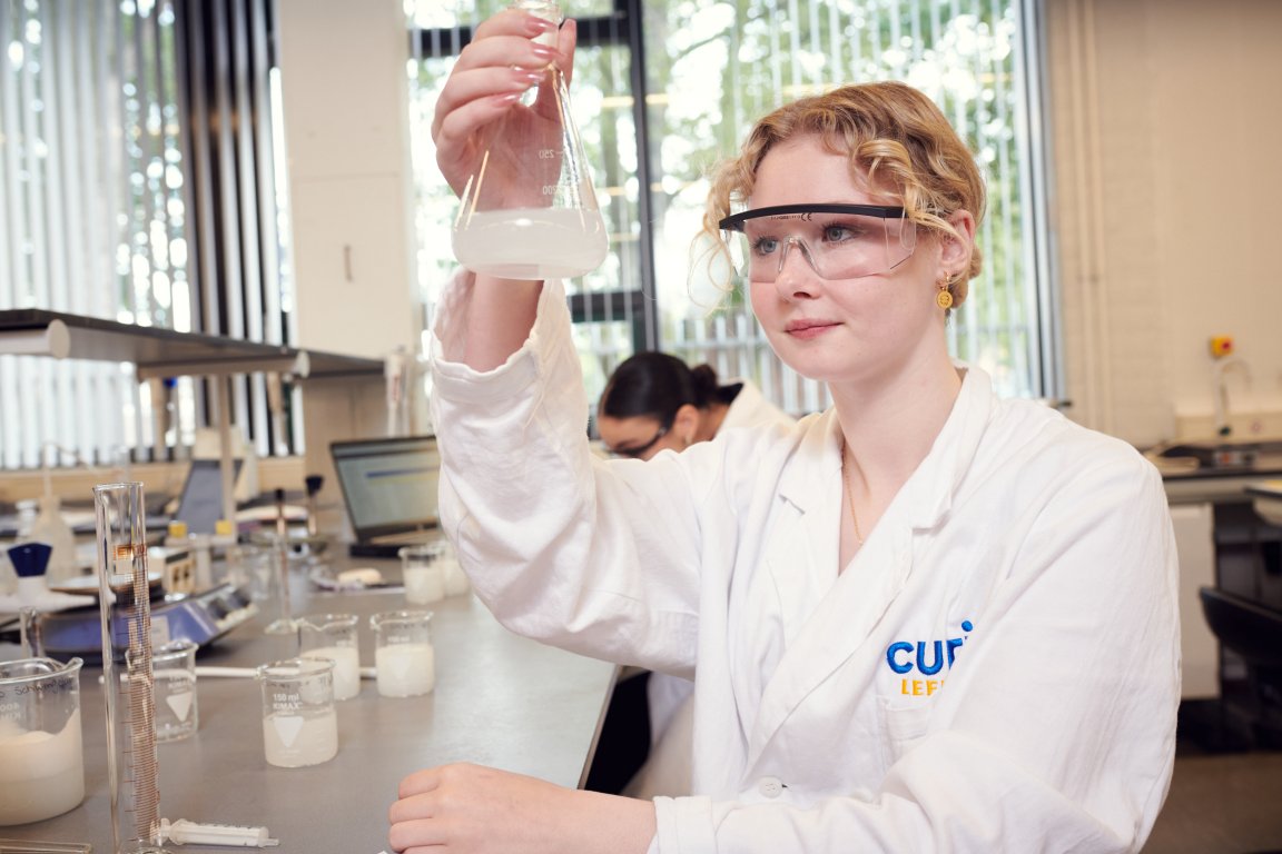 Studente in veiligheidskleding met een veiligheidsbril bekijkt een substantie in een kegelvormig glaasje