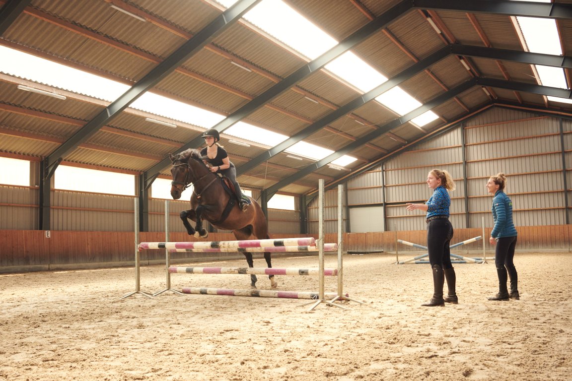 Drie studenten tijdens een les waarbij één student met een paard springt over een obstakel